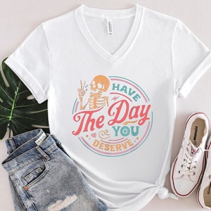 May include: White V-neck t-shirt with a colorful graphic of a skeleton with the text "Have The Day You Deserve". The shirt is paired with blue denim shorts and white sneakers.