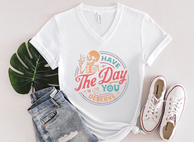 May include: White V-neck t-shirt with a colorful graphic of a skeleton with the text "Have The Day You Deserve". The shirt is paired with blue denim shorts and white sneakers.