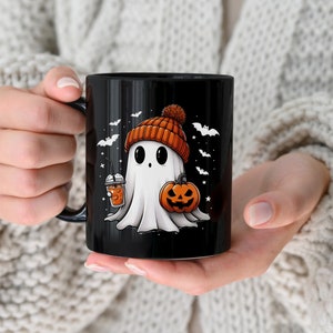 May include: Black ceramic mug with a white ghost wearing an orange and brown beanie holding a pumpkin and a cup of orange liquid. The ghost is surrounded by bats and stars.