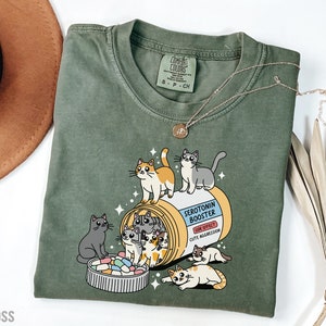 May include: Green t-shirt with a cartoon illustration of cats spilling out of a pill bottle labeled "Serotonin Booster". The text on the bottle reads "Side Effect: Cute Aggression".