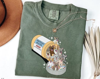 May include: Green t-shirt with a graphic of a pill bottle overflowing with cartoon dogs. The pill bottle says "Serotonin Booster" and "Side Effects: Cute Aggression". The text "Comfort Colors" is on a tag attached to the shirt.