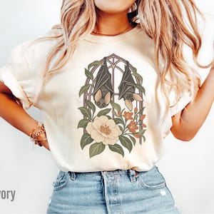 May include: An ivory short-sleeve t-shirt featuring a graphic print of two bats hanging inside a gothic-style arched window frame, surrounded by green leaves and large cream-colored flowers with small orange blossoms at the base.
