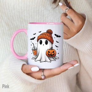 May include: A pink and white ceramic mug with a ghost wearing a hat and holding a pumpkin and a drink. The mug has a design of bats and stars.