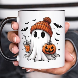 May include: White ceramic mug with a black handle. The mug features a cartoon ghost wearing an orange and brown knit hat, holding a pumpkin and a cup of orange juice. The background is white with black bats and stars.