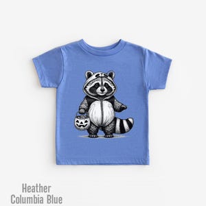 May include: A light blue t-shirt with a black and white illustration of a raccoon wearing a panda costume and holding a jack-o-lantern bucket. The raccoon is dressed for trick-or-treating.