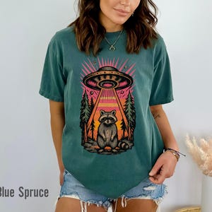 May include: Blue Spruce t-shirt featuring a graphic of a raccoon being beamed up by a UFO. The design includes a forest scene with a pink and orange sky. The text "Blue Spruce" is at the bottom.