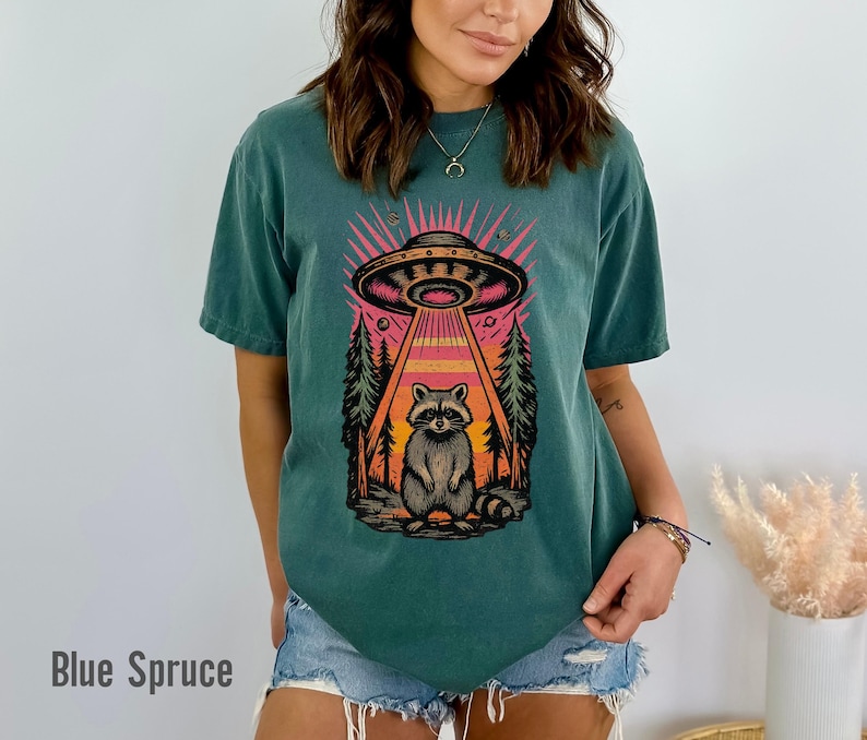 May include: Blue Spruce t-shirt featuring a graphic of a raccoon being beamed up by a UFO. The design includes a forest scene with a pink and orange sky. The text "Blue Spruce" is at the bottom.