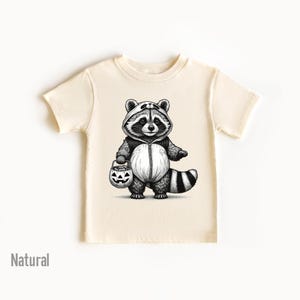 May include: A cream colored t-shirt with a black and white illustration of a raccoon wearing a costume and holding a jack-o-lantern.