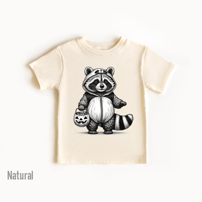 May include: A cream colored t-shirt with a black and white illustration of a raccoon wearing a costume and holding a jack-o-lantern.