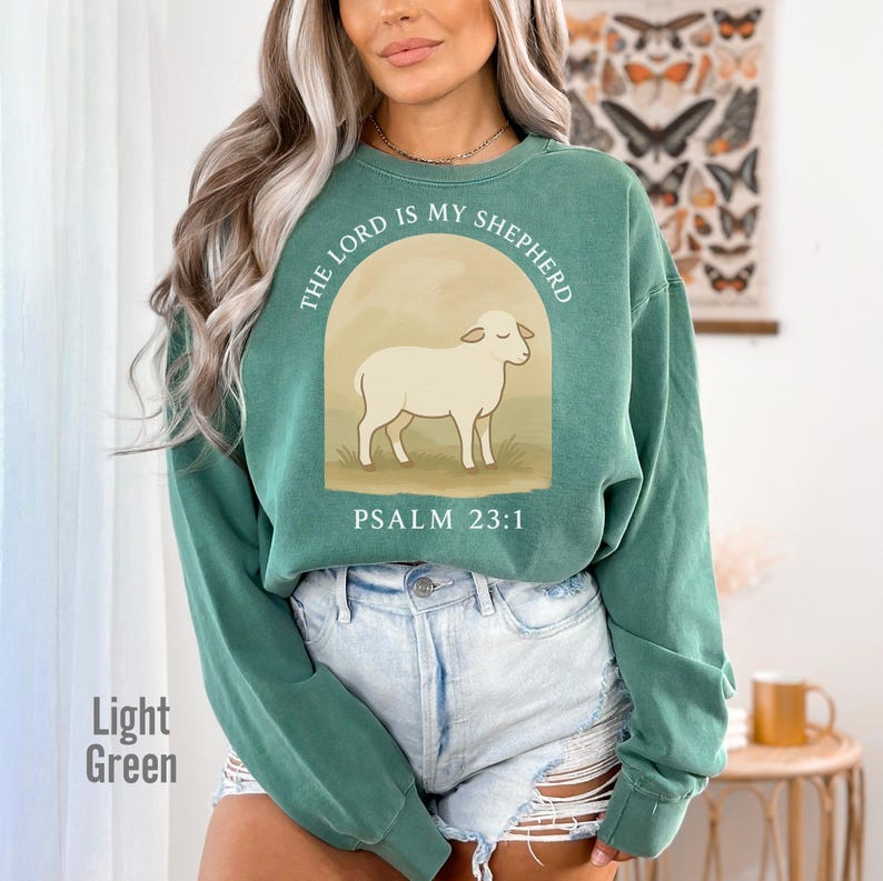 Op de afbeelding: Een lichtgroen sweatshirt met een witte schaap afbeelding en de tekst "The Lord is my Shepherd, Psalm 23:1".