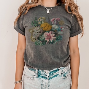 May include: A dark gray t-shirt with a floral print featuring a variety of flowers in shades of pink, yellow, and green. The shirt has a relaxed fit and rolled up sleeves.