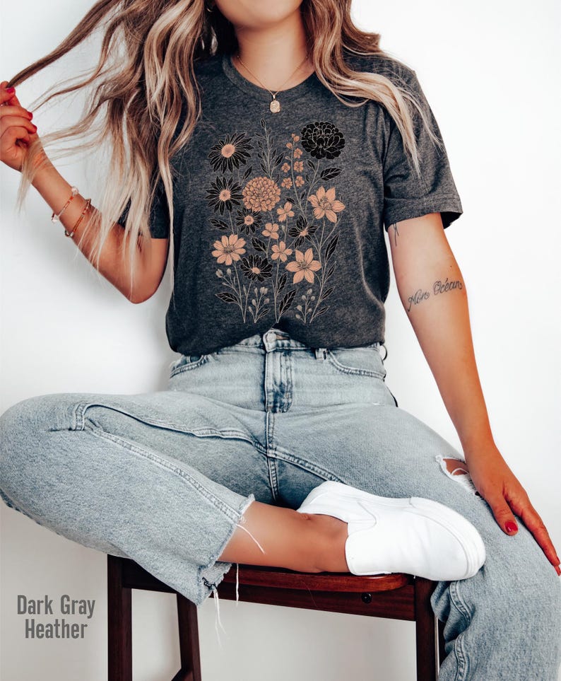 Op de afbeelding: Donkergrijze heather t-shirt met een bloemenontwerp in zwart en perziktinten. De afbeelding bevat verschillende bloemsoorten en stelen. Het shirt wordt gecombineerd met lichtblauwe jeans en witte sneakers. De tekst "Dark Gray Heather" is zichtbaar.