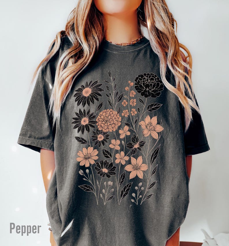 Peut inclure: T-shirt gris fonc&eacute; avec un motif floral en noir et p&ecirc;che. Le motif comprend diff&eacute;rents types de fleurs et de feuillage. Le mot "Pepper" est imprim&eacute; en bas &agrave; gauche du t-shirt.