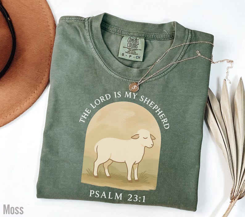 Op de afbeelding: Een groen T-shirt met een witte afbeelding van een schaap en de tekst "THE LORD IS MY SHEPHERD" en "PSALM 23:1".