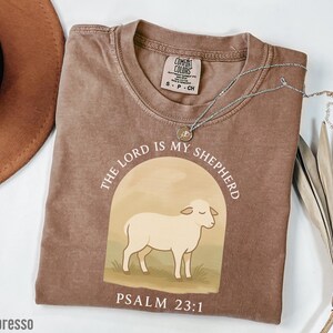 Op de afbeelding: Een bruin t-shirt met een witte afbeelding van een schaap en de tekst "THE LORD IS MY SHEPHERD" en "PSALM 23:1".