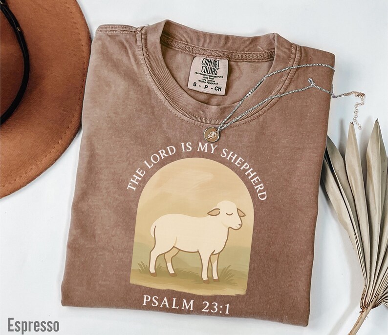 Op de afbeelding: Een bruin t-shirt met een witte afbeelding van een schaap en de tekst "THE LORD IS MY SHEPHERD" en "PSALM 23:1".