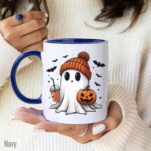 May include: A white ceramic mug with a blue rim features a cartoon ghost wearing an orange and brown knit hat. The ghost is holding a pumpkin and a cup of orange beverage with a straw. The background includes bats and stars.