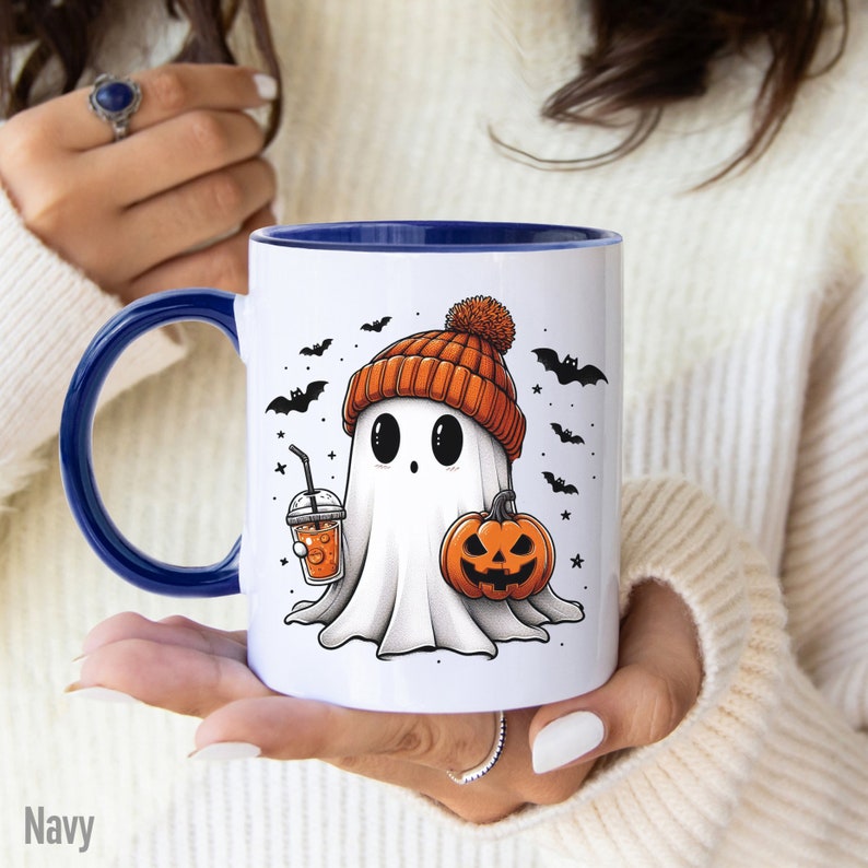 May include: A white ceramic mug with a blue rim features a cartoon ghost wearing an orange and brown knit hat. The ghost is holding a pumpkin and a cup of orange beverage with a straw. The background includes bats and stars.