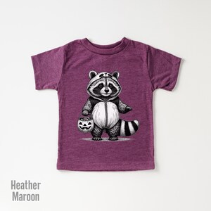 May include: A heather maroon t-shirt with a black and white illustration of a raccoon wearing a panda costume and holding a jack-o-lantern. The raccoon is dressed up for Halloween.