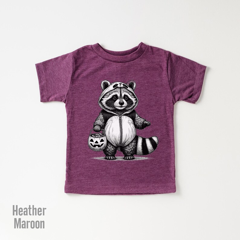 May include: A heather maroon t-shirt with a black and white illustration of a raccoon wearing a panda costume and holding a jack-o-lantern. The raccoon is dressed up for Halloween.