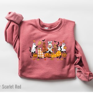 May include: Heather Scarlet Red sweatshirt featuring a Halloween-themed graphic. The design includes cartoon cats dressed as ghosts, scarecrows, and holding pumpkins. The cats are surrounded by pumpkins, leaves, and bats.