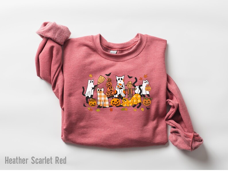 May include: Heather Scarlet Red sweatshirt featuring a Halloween-themed graphic. The design includes cartoon cats dressed as ghosts, scarecrows, and holding pumpkins. The cats are surrounded by pumpkins, leaves, and bats.