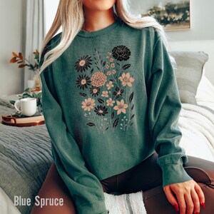 May include: A teal-colored sweatshirt featuring a floral design in black and peach. The graphic includes various flower types and foliage. The text "Blue Spruce" is visible at the bottom left of the image. The sweatshirt has long sleeves and a crew neck.