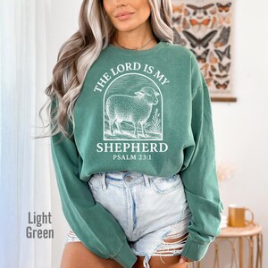 Puede incluir: Una sudadera de color verde claro con un gr&aacute;fico blanco de una oveja y el texto "The Lord is my Shepherd, Psalm 23:1".