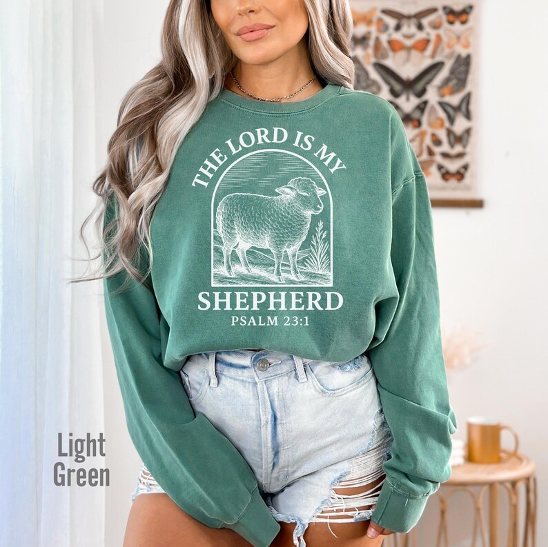 Puede incluir: Una sudadera de color verde claro con un gr&aacute;fico blanco de una oveja y el texto "The Lord is my Shepherd, Psalm 23:1".