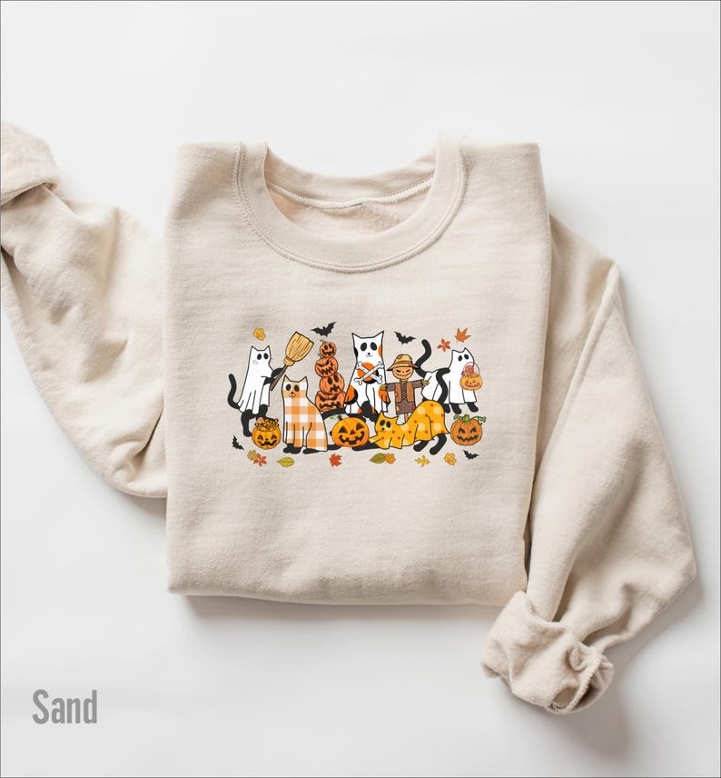 May include: Sand-colored sweatshirt featuring a Halloween-themed graphic. The graphic includes cartoon cats dressed as ghosts, pumpkins, and scarecrows. The word "Sand" is printed in the lower left corner.