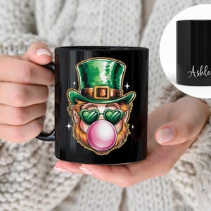 Könnte beinhalten: Schwarze Keramiktasse mit einem Grafikdruck eines Cartoon-Kobolds mit grüner Sonnenbrille und grünem Hut, der eine rosa Blase bläst. Die Tasse trägt den Namen "Ashley".