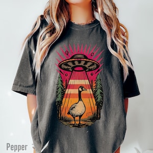 May include: Dark gray t-shirt featuring a graphic of a goose standing under a UFO with a beam of light. The design includes a retro sunset and pine trees. The word "Pepper" is printed at the bottom left.