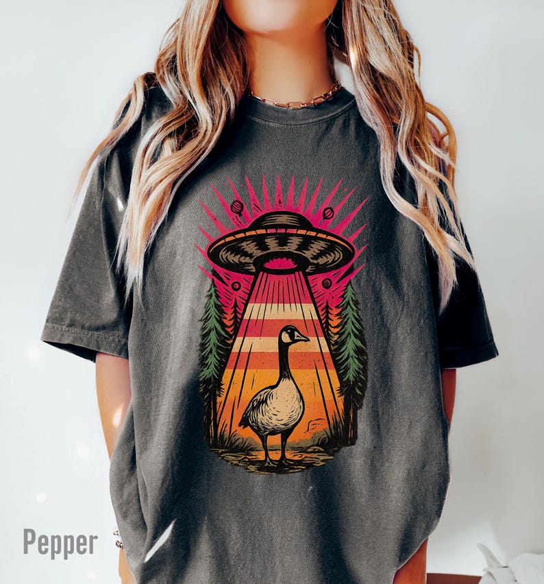 May include: Dark gray t-shirt featuring a graphic of a goose standing under a UFO with a beam of light. The design includes a retro sunset and pine trees. The word "Pepper" is printed at the bottom left.