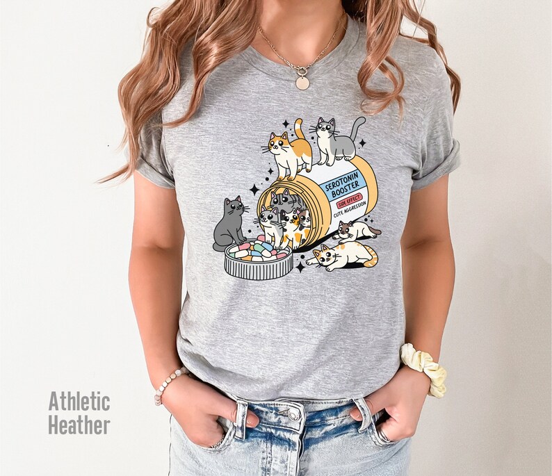May include: Gray t-shirt with a graphic of a pill bottle labeled "Serotonin Booster" with cute cartoon cats spilling out of it.