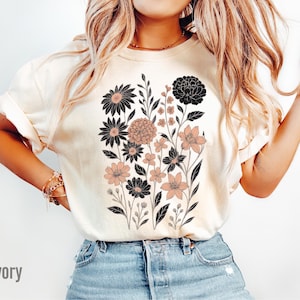 Peut inclure: T-shirt ivoire avec un motif floral noir et p&ecirc;che. Le graphisme comprend divers types de fleurs et de feuillage. Le t-shirt est de coupe d&eacute;contract&eacute;e et a des manches courtes. Le mot "Ivory" est imprim&eacute; en bas &agrave; gauche.