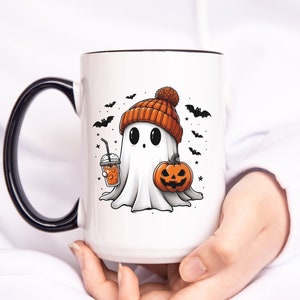 May include: White ceramic mug with a black rim and handle. The mug features a cartoon ghost wearing an orange and brown knit hat, holding a pumpkin and a cup of orange juice. The background is white with black bats and stars.
