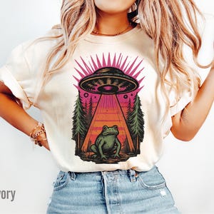 May include: Ivory-colored t-shirt featuring a graphic of a UFO beaming a frog. The design includes a pink and black UFO, a frog, and a forest scene. The word "Ivory" is printed at the bottom left.