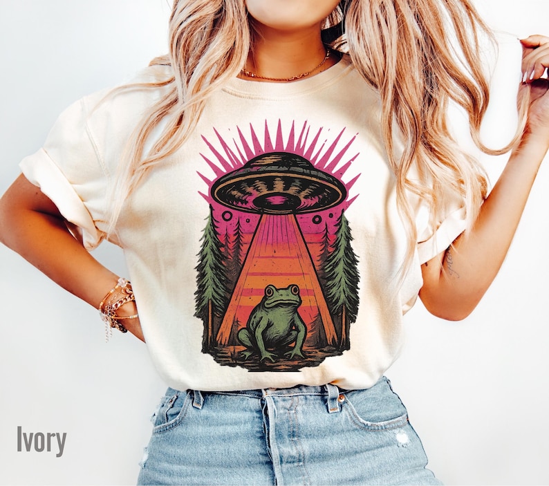 May include: Ivory-colored t-shirt featuring a graphic of a UFO beaming a frog. The design includes a pink and black UFO, a frog, and a forest scene. The word "Ivory" is printed at the bottom left.