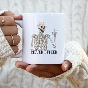 Op de afbeelding: Witte keramische mok met een zwart-witte illustratie van een skelet dat een ok&eacute;-teken geeft. De tekst "NEVER BETTER" is onder het skelet gedrukt.