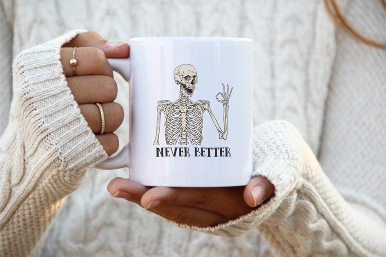 Op de afbeelding: Witte keramische mok met een zwart-witte illustratie van een skelet dat een ok&eacute;-teken geeft. De tekst "NEVER BETTER" is onder het skelet gedrukt.