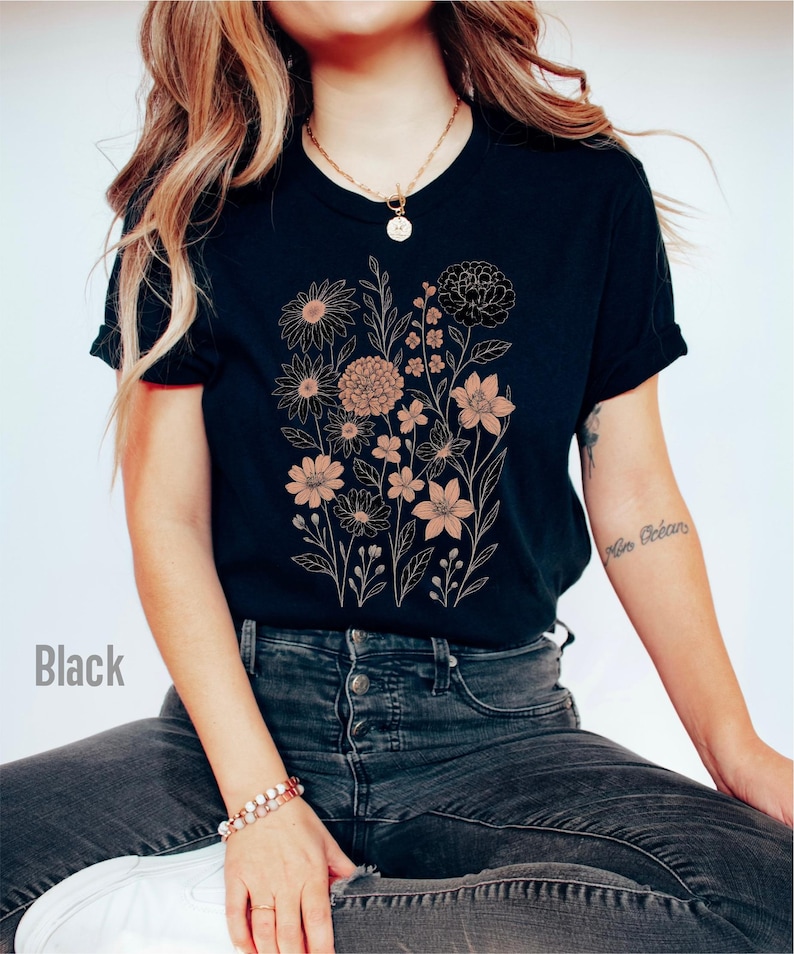Op de afbeelding: Zwart t-shirt met een bloemenontwerp in perzik- en goudtinten. Het ontwerp bevat verschillende bloemen en bladeren. Het woord "Black" staat linksonder in de afbeelding.
