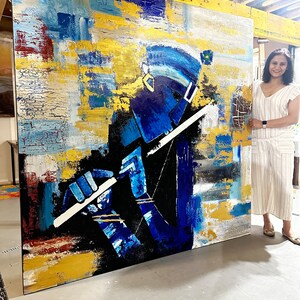 May include: Abstract painting of a person playing a flute, with a blue, gold, and white colour scheme. The painting is large and is leaning against a wall.