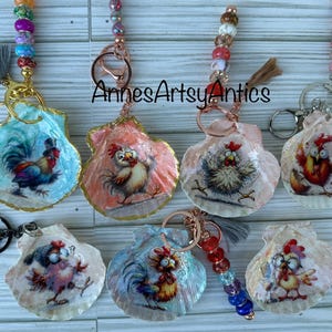 May include: Hand-painted seashell keychains featuring rooster and chicken designs. Each shell is adorned with a unique illustration and accented with colorful beads and a tassel. The keychains are attached to metal rings.