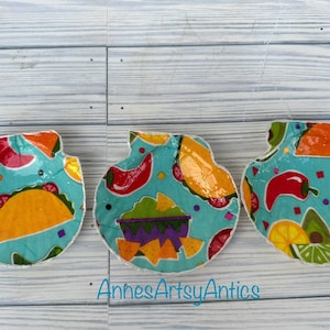 May include: Three turquoise blue, white, and yellow fabric ornaments with a Mexican food theme. The ornaments feature images of tacos, guacamole, and peppers.