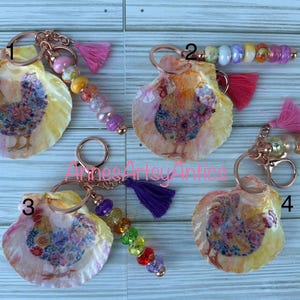 May include: Four seashell keychains with a painted hen design. Each keychain has a different colored tassel and beaded accent. The keychains are attached to a gold-colored ring.