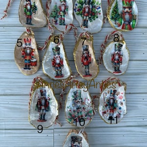 May include: A collection of twelve Christmas ornaments. Each ornament features a nutcracker design on a white oyster shell with a gold-painted edge. The ornaments are strung with red and gold ribbon for hanging.