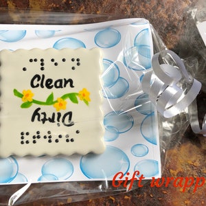 May include: A white ceramic tile with a blue and white bubble pattern background. The tile has a white border and says "Clean" in black lettering with a green vine and yellow flowers. The tile also says "Dirty" in black lettering with braille dots. The tile is wrapped in clear plastic with a white ribbon.