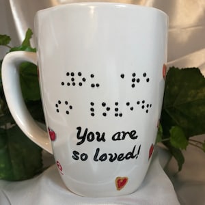 May include: White ceramic mug with black dots spelling "You are so loved!" in braille. The mug is decorated with red hearts.