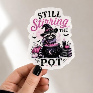 May include: A die-cut sticker featuring a raccoon wearing a witch hat and stirring a black cauldron filled with pink liquid. The text reads STILL STIRRING THE POT. The design includes pink pumpkins, a stack of books labeled SPELLS POTIONS & CHAOS, and bats.