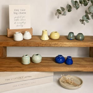 May include: A collection of small ceramic vases in various shapes and colors, including white, yellow, blue, and green, displayed on wooden shelves. A sign reads "tiny vase big charm perfect for little stems & everyday beauty."